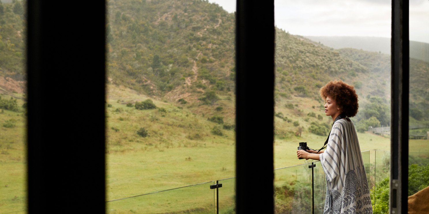 Side view of young Afro woman with binoculars standing in balcony seen from rental villa's window