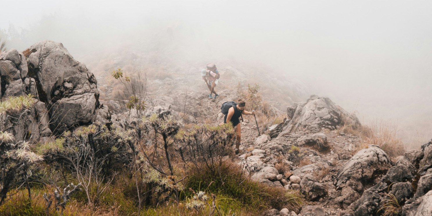Hikers climbing a rocky mountain trail in thick fog, surrounded by rugged terrain and sparse vegetation. Hikers climbing a rocky mountain trail in thick fog, surrounded by rugged terrain and sparse vegetation.