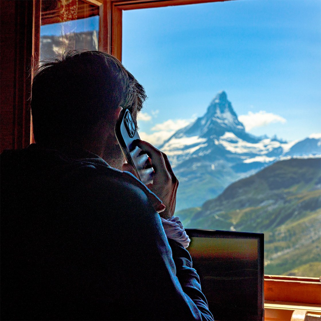 Man on a phone call while working on a laptop, looking out of a window at the Matterhorn mountain in Switzerland. Man on a phone call while working on a laptop, looking out of a window at the Matterhorn mountain in Switzerland.