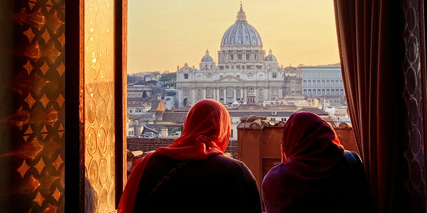 Two people in red headscarves looking out of a window at St. Peter’s Basilica in Vatican City during sunset