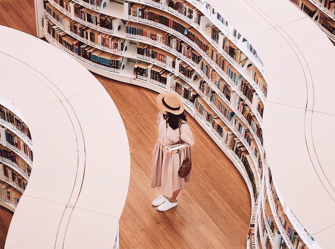 Woman in a pink dress and straw hat standing among curved shelves filled with books in a modern library. Woman in a pink dress and straw hat standing among curved shelves filled with books in a modern library.