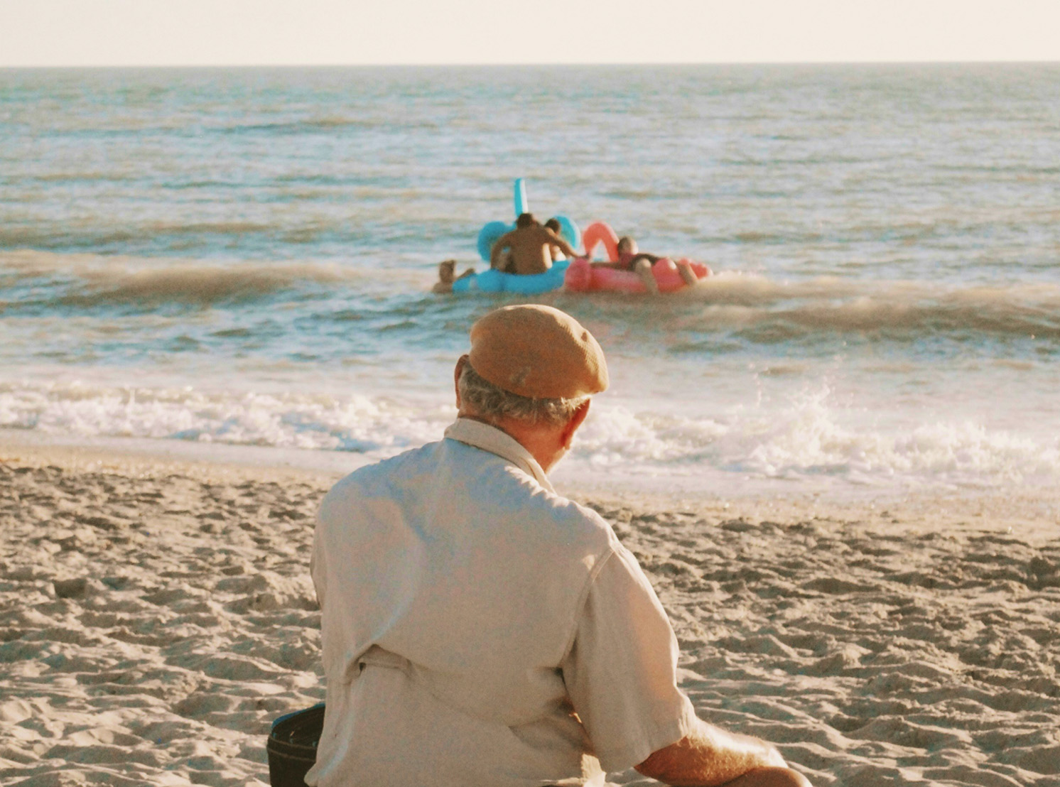 Elderly man sitting on the sand watching children play with inflatable floats in the sea. Elderly man sitting on the sand watching children play with inflatable floats in the sea.