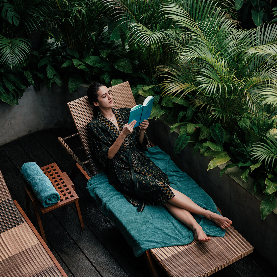 Woman relaxing on a lounge chair surrounded by lush tropical plants, reading a book in a peaceful spa-like setting. Woman relaxing on a lounge chair surrounded by lush tropical plants, reading a book in a peaceful spa-like setting.